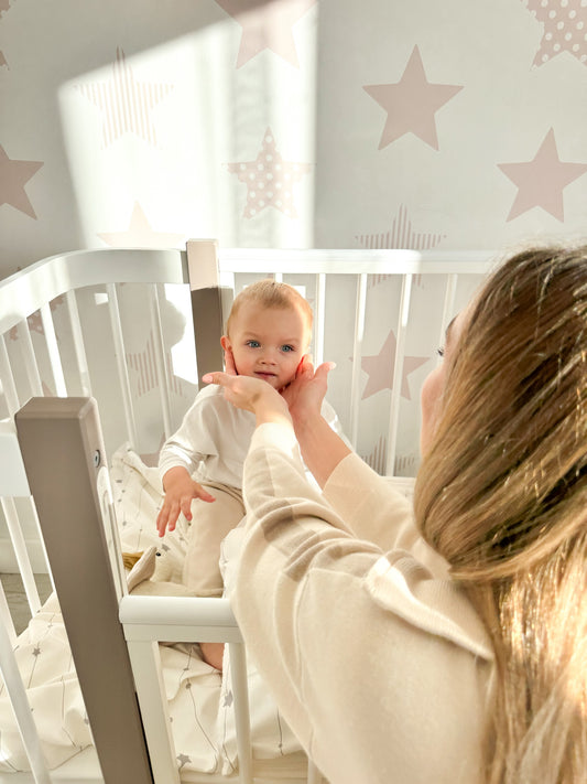 Happy mother leaning over the ELIN baby crib to kiss her newborn baby in a sunlit nursery