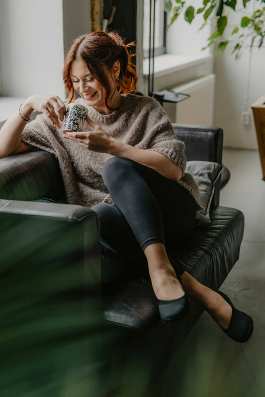 Cozy indoor lifestyle shot of a model relaxing cross-legged on a couch while sipping tea, wearing the comfortable Upcycled Ballerina Shoes made from sustainable recycled materials.