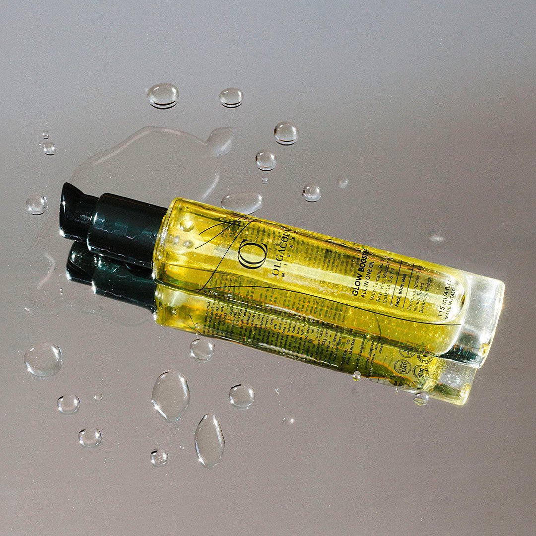 Olgacola Milano Glow Booster All In One Oil in a glass bottle resting on a reflective silver surface with water droplets. A luxury botanical oil for face, body, and hair.