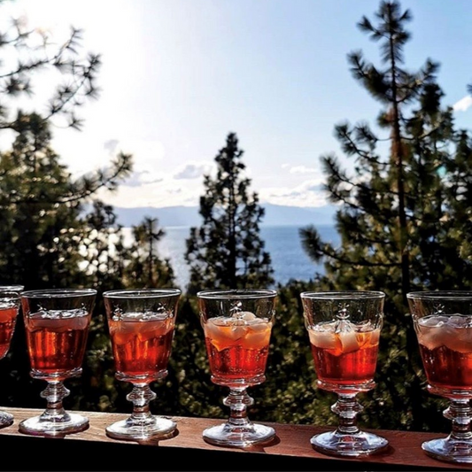 A lifestyle photograph of multiple La Rochere Bee glasses filled with ice and drinks (on the rocks) on a terrace, with a breathtaking mountain range and clear sky in the background.