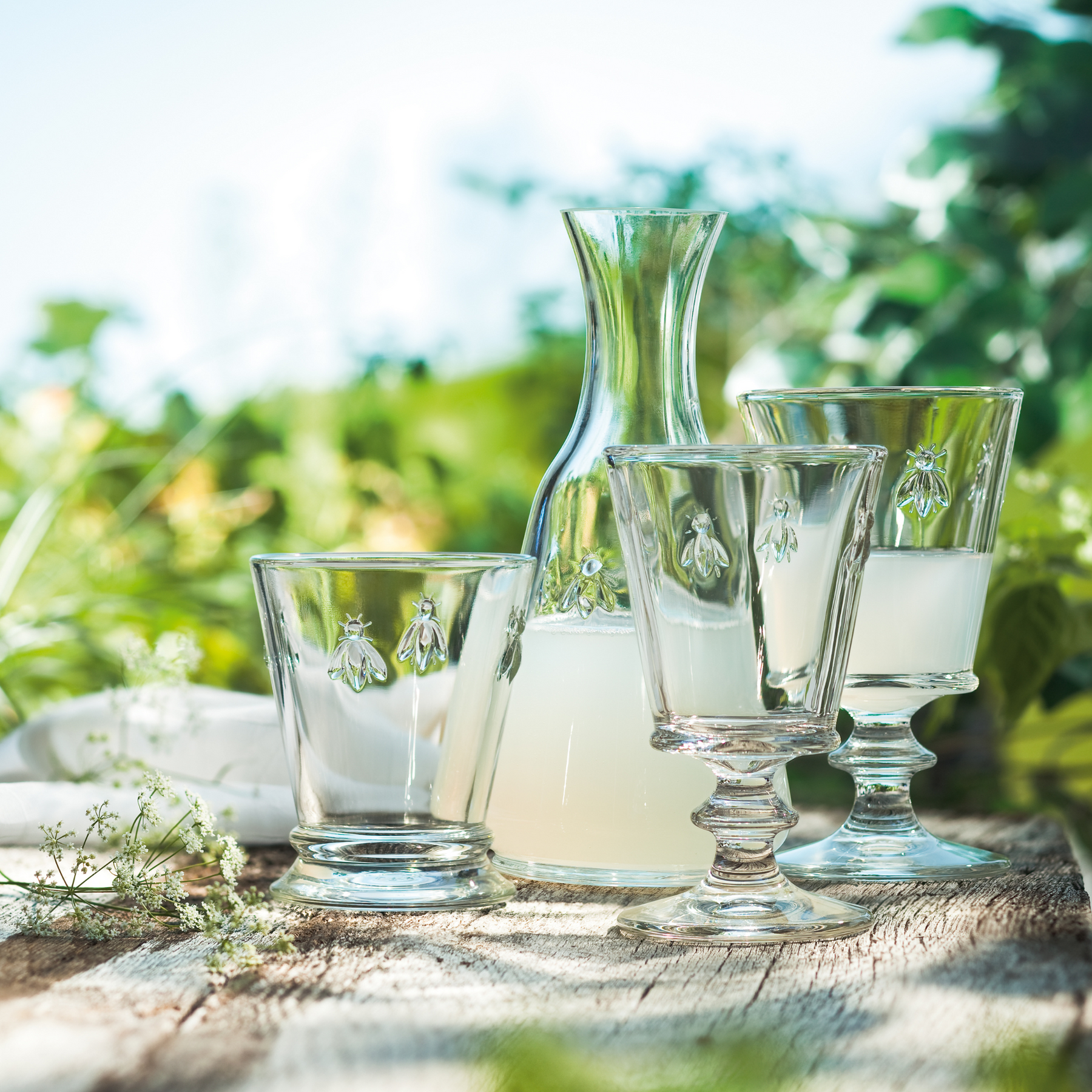 A rustic outdoor scene featuring a single La Rochere Bee glass on an aged wooden plank, grouped with other matching La Rochere glassware and products.