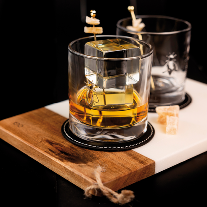 Clear La Rochère Bee whiskey glass filled with amber whiskey and ice cubes, resting on a small wooden serving board