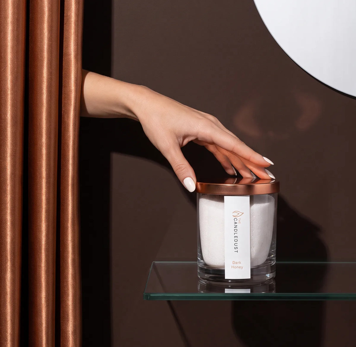 Woman's hand reaching from behind an elegant curtain to touch a 160g Dark Honey scented glass candle on a glass shelf