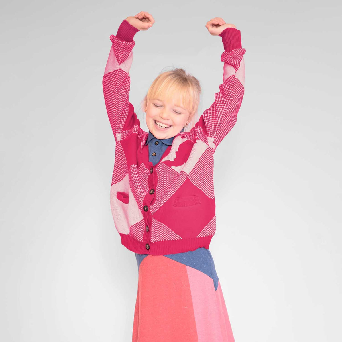 Child wearing Infantium Victoria pink and white organic cotton knitted cardigan with arms raised against a light gray background