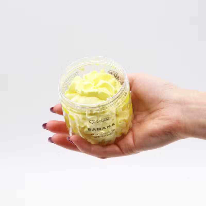 Woman's hand holding an open jar of banana whipped soap, displaying its fluffy yellow swirled texture
