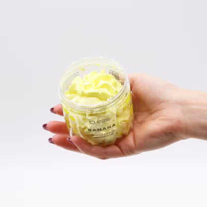 Woman's hand holding an open jar of banana whipped soap, displaying its fluffy yellow swirled texture