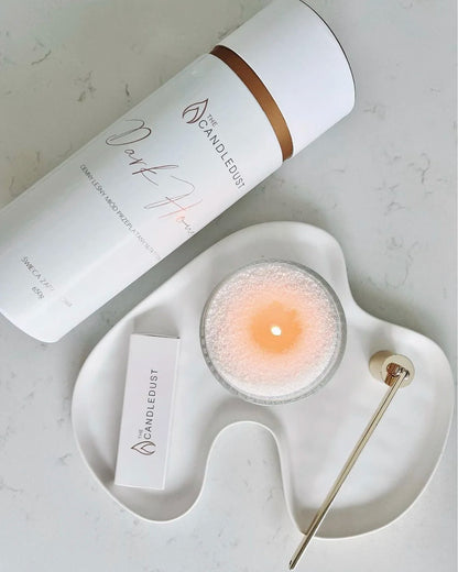 Top view of a 650g Candledust Dark Honey tube and a lit pearled candle burning in a glass jar on a modern tray