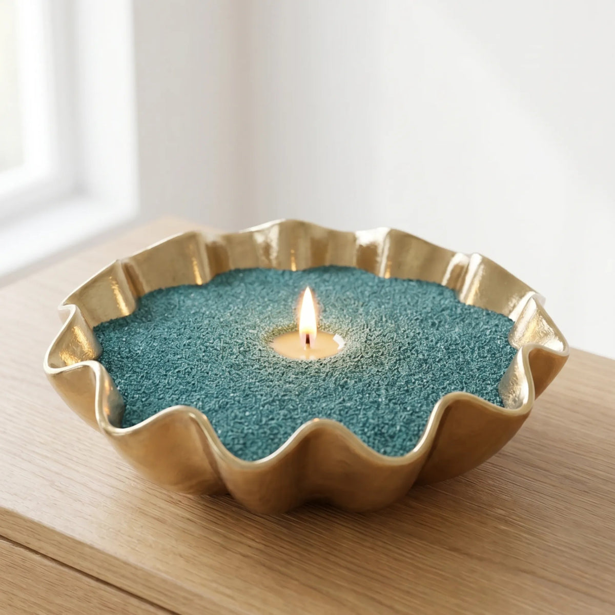 A gold-toned scalloped metal candle holder filled with teal-green sand, holding a lit tealite candle on a wooden surface.