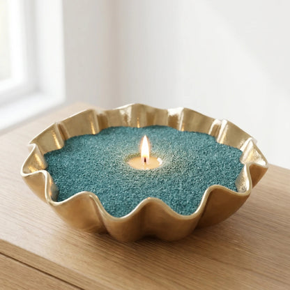 A gold-toned scalloped metal candle holder filled with teal-green sand, holding a lit tealite candle on a wooden surface.
