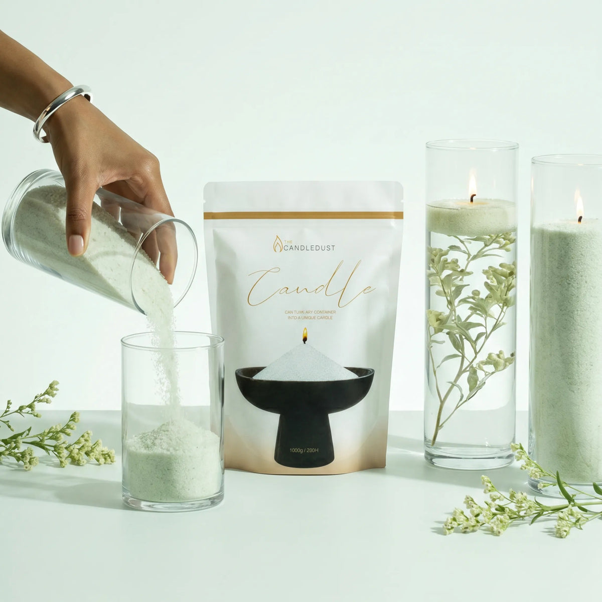Woman pouring Lemongrass Ginger pearled candle sand into a glass holder on a white table next to two burning sand candles