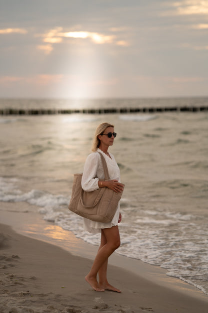 Moi Mili Mom Weekender Bag Latte: a mother walking along the shore with a sturdy quilted bag on her shoulder at sunset