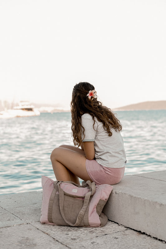 Moi Mili Mom Weekender Bag Pink Coffee: back view of a young woman sitting on a dock with her large quilted cotton bag beside her at the waterfront