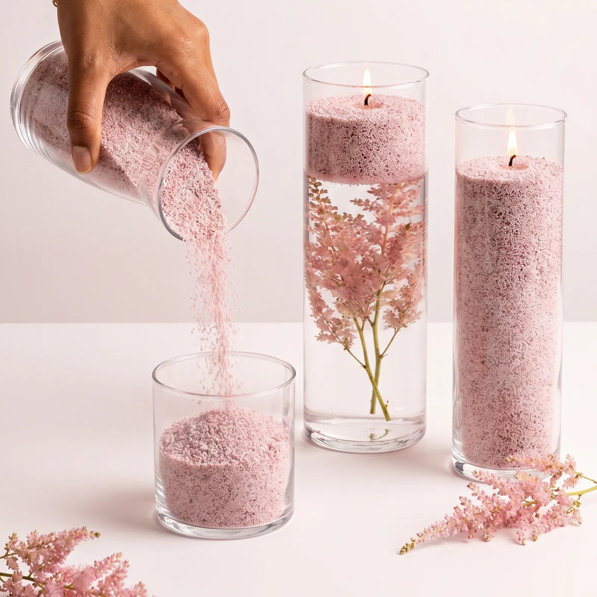 Woman pouring Nude pearled candle sand into a glass holder on a white table next to two burning sand candles