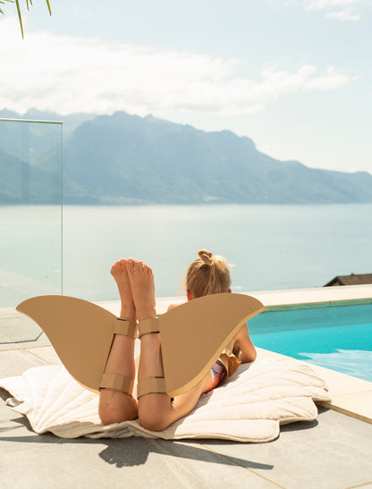 Moi Mili Beige Shell Playmat: a toddler girl resting on a plush velvet shell-shaped baby mat at a poolside with a sea and mountain view