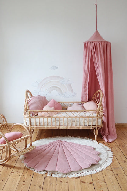 Dreamy nursery decor: a natural wooden baby bed with a hanging canopy, a vintage rocking chair, and a Moi Mili Powder Pink Velvet Shell Playmat on the floor