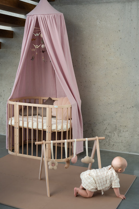 Baby playtime: a newborn exploring wooden toys on a quilted floor mat, sheltered under a Moi Mili Baby Pink Organic Cotton Canopy in a cosy nursery