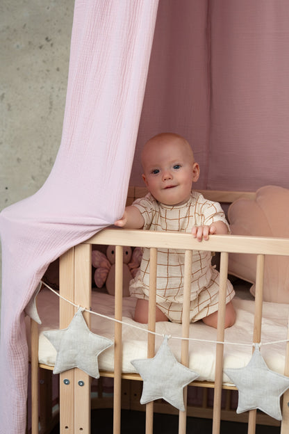 Baby playing in a crib, looking up at the soft hanging folds of a Moi Mili Baby Pink Organic Cotton Canopy in a bright nursery