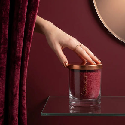 Woman's hand reaching from behind an elegant sangria red curtain to touch a 160g Sangria Red unscented glass candle on a glass shelf