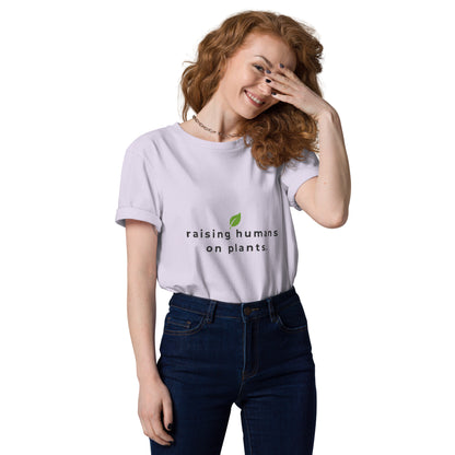 Smiling model wearing an eco-friendly lavender t-shirt. On the chest is printed the phrase "raising humans on plants." in charcoal grey with a small leaf centered above the text.